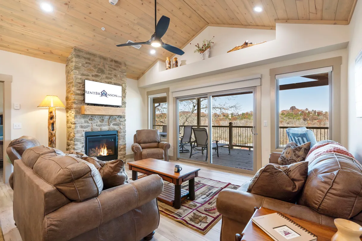 The image showcases a warm, inviting living room featuring a striking stone fireplace, soaring wooden ceilings, large sunlit windows that offer glimpses of the outdoors, and plush seating arranged for comfort and togetherness. The overall atmosphere is cozy and welcoming, perfect for gathering with loved ones.