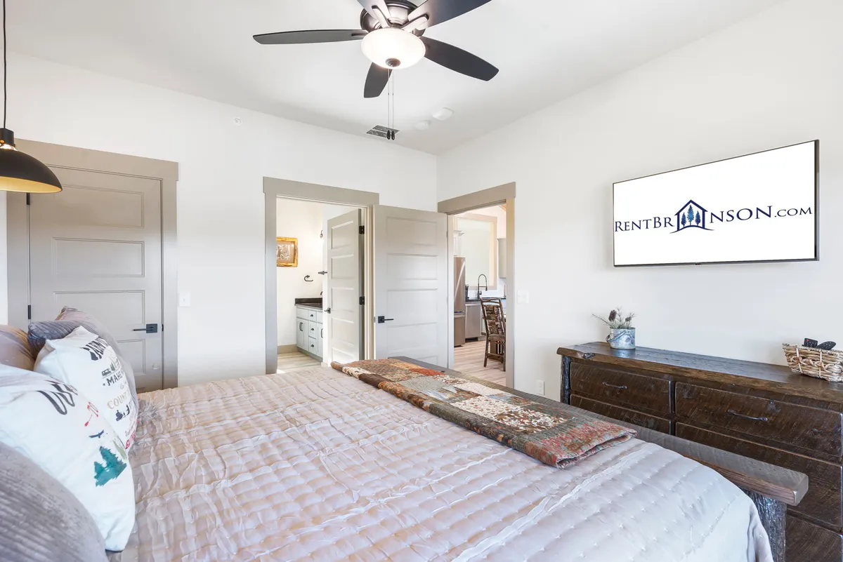 A serene king-size bedroom featuring a spacious bed adorned with decorative pillows, a wooden dresser, and a wall-mounted television. The room is bathed in natural light, creating a comfortable, restful atmosphere ideal for unwinding.