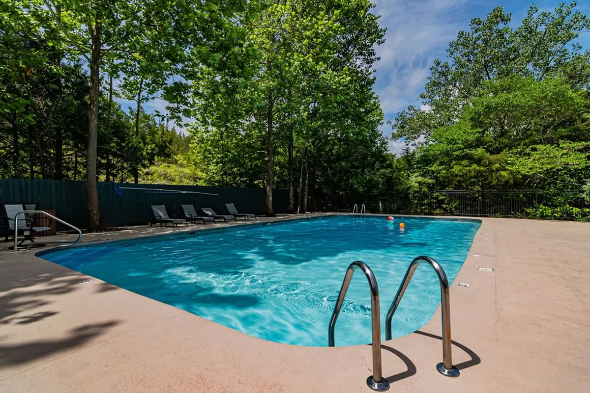 An outdoor swimming pool set among trees, bordered by lounge chairs, suggesting a relaxed, recreational environment. The area appears welcoming for families, offering a space to swim, sunbathe, or socialize, with greenery providing a calming backdrop.