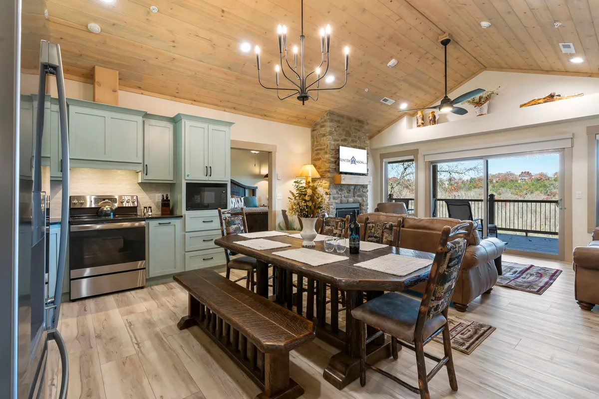 A wide-angle view of the open-concept area showcasing a warm stone fireplace, a comfortable living room with plush seating, a large dining table, a bright kitchen island, and large farmhouse windows letting in natural light. The scene captures multiple zones—kitchen, dining, and living—within the same space, emphasizing flow and togetherness.