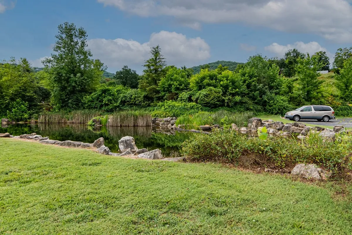 Featuring a calm water surface backed by lush greenery, natural rock formations, and distant hills, this view exudes a sense of peaceful escape. A parked vehicle hints at easy access for day trips and exploration.