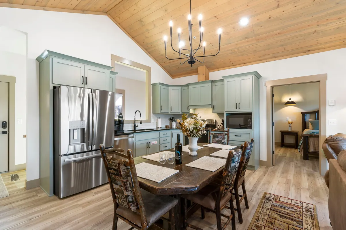 A close-up perspective of the dining and kitchen area with farmhouse-style cabinetry, a wooden dining table with comfortable chairs, a chandelier overhead, and stainless steel appliances. The vibe is cozy, warm, and inviting, perfect for meals and social gatherings.